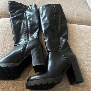 Svarta höga stövlar med klack - Snygga svarta stövlar i skinnimitation med grov sula och hög blockklack. Dragkedja på insidan för enkel påtagning och en stilren, minimalistisk look. Perfekta för dig som vill ha en edgy vibe till din outfit.