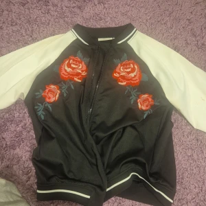 Svart bomberjacka med broderade rosor - Snygg svart bomberjacka från KappAhl med vita ärmar och broderade röda rosor framtill. Jackan har dragkedja och sportiga ränder vid muddar och krage. Perfekt för dig som vill sticka ut med coola detaljer och blommigt broderi.