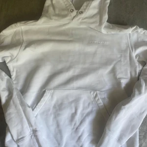 Vit hoodie från J.Lindeberg - Vit hoodie från J.Lindeberg med broderad logga på bröstet. Klassisk modell med huva, känguruficka och långa ärmar. Tillverkad i mjuk bomull, perfekt för en clean och stilren look. Skick 8/10 pris kan diskuteras, passar L 