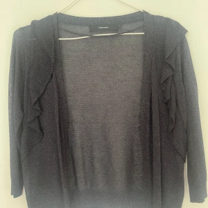 Mörkgrå kofta med volang från Vero Moda - Snygg mörkgrå kofta från Vero Moda i tunt, stickat material. Koftan har långa ärmar och volangdetaljer längs framsidan som ger en extra touch. Perfekt att slänga över en topp för en chill och stilren look.