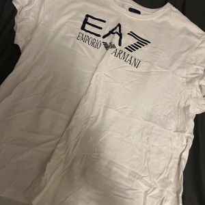 Vit EA7 Emporio Armani t-shirt - Snygg vit t-shirt från EA7 Emporio Armani med stor svart logga och tryck framtill. Klassisk rund hals och korta ärmar. Perfekt för dig som gillar stilrena och exklusiva plagg med tydlig branding.