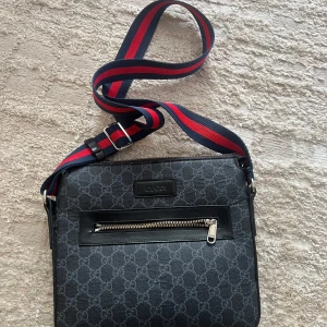 Svart axelväska från Gucci med GG-mönster - Snygg svart axelväska från Gucci med klassiskt GG-mönster över hela väskan. Väskan har en bred, justerbar axelrem i rött och marinblått, samt detaljer i svart skinn. Framsidan har en dragkedjeficka och diskret Gucci-logga i svart läder.