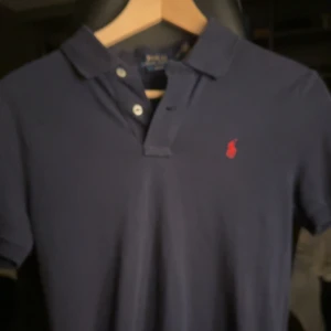 Mörkblå pikétröja från Ralph Lauren - Säljer en mörkblå pikétröja från Polo Ralph Lauren i slim fit. Tröjan har klassisk krage, två knappar och den ikoniska röda loggan broderad på bröstet. Perfekt för dig som gillar stilren och tidlös design.
