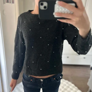 Mörkgrå stickad tröja med pärlor - Mysig mörkgrå stickad tröja med rund hals och långa ärmar. Tröjan är dekorerad med små pärlor över hela plagget för en extra snygg touch. Perfekt för dig som gillar en enkel men ändå trendig look.