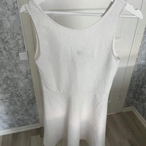 Vit klänning från Divided - Snygg vit klänning från Divided med lätt struktur i tyget. Modellen är ärmlös med rundad halsringning fram och v-ringning bak. Klänningen har en kort längd och är perfekt för sommarens alla tillfällen. Enkel och stilren design som är lätt att matcha. Vid behov av fler bilder eller frågor hör av dig!