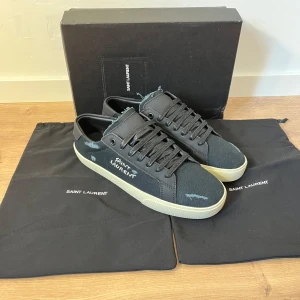 Saint Laurent sneakers - Säljer ett par ÄKTA svarta sneakers från Saint Laurent med handskriven logga på sidan. Skorna har rund tå, platt sula och är tillverkade i canvas med svarta snören. Klassisk och stilren design med diskreta detaljer. Storlek 41 sitter som 41. Skick 10/10. Tillbehör/OG låda och dustbags.