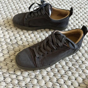 Christian Louboutin svarta sneakers mocka - Snygga svarta sneakers från Christian Louboutin i mjuk mocka med klassisk röd sula och rund tå. Skorna har snörning och en stilren design med diskreta detaljer och beige innersida. Perfekta för dig som vill ha en lyxig touch på din outfit.