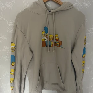 Beige Simpsons hoodie med tryck - Säljer en beige hoodie med stort Simpsons-tryck på bröstet där hela familjen sitter i soffan. Ärmarna har coola tryck med ansikten på Homer, Marge, Bart, Lisa och Maggie. Tröjan har huva med snörning och en stor magficka. Perfekt för dig som älskar serien!