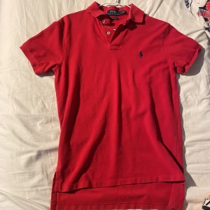 Röd pikétröja från Polo Ralph Lauren - Klassisk röd pikétröja från Polo Ralph Lauren med broderad logga på bröstet. Tröjan har korta ärmar, krage och knappar framtill. Custom fit-modell i mjuk bomull som är skön att bära och passar perfekt till jeans eller shorts.