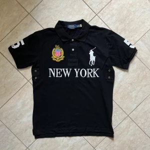 Svart Polo Ralph Lauren New York t-shirt - Svart t-shirt från Polo Ralph Lauren med broderad logga, stort vitt tryck 'NEW YORK' och emblem på bröstet. Klassisk pikékrage, korta ärmar och stora siffror på ärmarna och ryggen. Tillverkad i bomull, perfekt för en sportig och stilren look.
