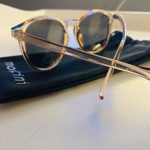 Runda solglasögon från Montini - Snygga runda solglasögon från Montini med transparent beige plastbåge och mörka linser. Kommer med ett svart fodral med Montini-logga. Perfekta för att lyfta din stil med en cool och modern touch.