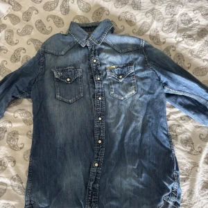 Blå jeansskjorta från Replay - Snygg blå jeansskjorta från Replay med klassisk krage, två bröstfickor med knappar och vita tryckknappar längs hela framsidan. Skjortan har lång ärm och coola slitningar för en avslappnad look. Perfekt till jeans eller svarta byxor.