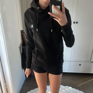 Svart hoodie med snörning och huva - Svart hoodie med klassisk huva och snörning, samt en liten broderad detalj framtill. Tröjan har en stor magficka och långa ärmar med muddar. Perfekt för chill dagar och enkel att matcha med allt. Mjuk bomullskänsla och stilren look.