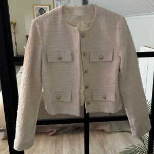 Beige kavajjacka från H&M, storlek S - Stilren beige kavajjacka från H&M i storlek S. Jackan är tillverkad i ett strukturerat tyg med fyra fickor framtill och snygga guldknappar. Rak passform, rund halsringning och helfodrad insida.