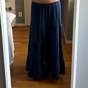 Mörkblå långkjol med volanger - Säljer en mörkblå långkjol med flera volangpartier och resår i midjan. Kjolen är luftig och har en avslappnad passform, perfekt för dig som gillar en bohemisk vibe. Materialet känns mjukt och flowy, vilket gör den superbekväm att bära hela dagen.