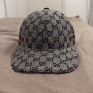 Gucci keps navy  - Snygg keps från Gucci i beige och svart med klassiskt GG-monogram över hela kepsen. På sidan finns en ikonisk röd och marinblå rand. Kepsen är tillverkad i bomull och har en böjd skärm. Perfekt för dig som vill sticka ut med en lyxig accessoar.