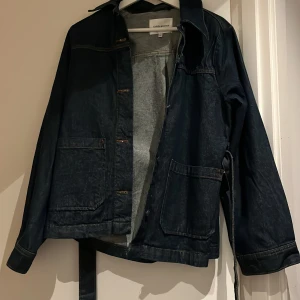Mörkblå jeansjacka med bälte Carin Wester - Snygg mörkblå jeansjacka från Carin Wester i klassisk modell med stora fickor framtill och bälte i midjan. Jackan har långa ärmar, krage och knäppning med knappar. Den är köpt förra hösten och är använd några fåtal gånger🩷