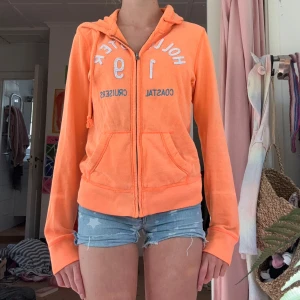 Orange ziphoodie från Hollister - Säljer en orange ziphoodie från Hollister med broderad text och siffror framtill. Jackan har huva med dragsko, två fickor och dragkedja hela vägen. Perfekt för chill dagar och snygg till jeansshorts eller leggings.