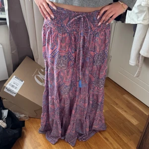Lång paisleymönstrad kjol med snörning - Flowig långkjol i rosa och lila med paisleyinspirerat mönster. Kjolen har smock i midjan och justerbar snörning med tofsdetalj. Tofsarna som hänger på trådarna e lite trasiga och en saknas. Köpt i Danmark för några år sen vet inte märket. 