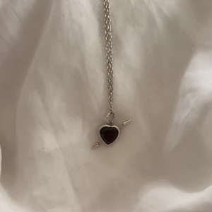 Maria Nilsdotter Halsband  - Snyggt halsband i silverfärg med ett rött hjärthänge genomborrat av en pil. 