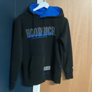 Svart Hoodrich hoodie med blå detaljer - Svart hoodie från Hoodrich med blått foder i huvan och blå logga framtill och baktill. Tröjan har snörning i huvan, känguruficka och tryck på ärmen. Perfekt för dig som gillar streetwear och vill sticka ut med coola detaljer.