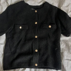 Svart stickad kofta från Gina Tricot - Svart stickad kofta från Gina Tricot med korta ärmar och snygga guldknappar framtill. Två dekorativa fickor på bröstet och ribbade kanter ger en trendig look. Perfekt att slänga över en topp för en chill men stilsäker vibe.