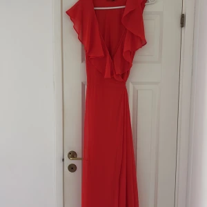 Röd långklänning med volang från veromoda - Säljer en snygg röd långklänning med djup v-ringning och volangdetaljer upptill. Klänningen har omlottkänsla och knyt i midjan för en smickrande siluett. Perfekt för dig som vill sticka ut med en färgstark look!