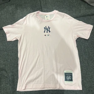 Ljusrosa New York Yankees t-shirt Champion - Ljusrosa t-shirt från Champion med New York Yankees-logga broderad på bröstet och officiell patch nertill. Klassisk rund hals och korta ärmar. Skön bomullskvalitet och relaxed fit, perfekt för dig som gillar sportiga vibes och streetwear.