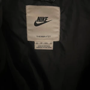 Svart pufferjacka Nike Therma-Fit XS - Säljer en svart pufferjacka från Nike i storlek XS men kan upplevas stor i storlek.Jackan har Therma-Fit-teknologi, huva med snörning och dragkedja framtill. Snyggt broderad Nike-logga på bröstet och glansig finish. Perfekt för kalla dagar när du vill hålla stilen.