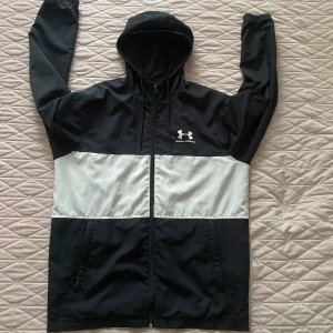 Svart och vit vindjacka Under Armour - Snygg svart och vit vindjacka från Under Armour med huva och dragkedja hela vägen. Jackan har meshfoder, två fickor och coolt logotryck på bröstet. Perfekt för dig som gillar sportig stil och vill ha något lätt och funktionellt.