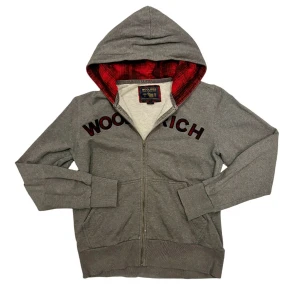 Grå hoodie från Woolrich med röd huva - Snygg grå hoodie från Woolrich med dragkedja och stor huva fodrad i rött rutigt tyg. Stora svarta och röda bokstäver över bröstet. Mjuk bomullskänsla, två fickor fram och ribbade muddar. Perfekt för chill dagar eller när du vill ha en avslappnad look. Det är en L men sitter exakt som en M 👍🏼