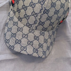 Gucci keps i merinoull och kashmir - Exklusiv keps från Gucci i beige och marinblått med klassiskt GG-mönster över hela kepsen. Tillverkad i 90% merinoull och 10% kashmir. Snygga röda och gröna detaljer på sidorna. Perfekt accessoar för dig som vill sticka ut med lyxig streetstyle.