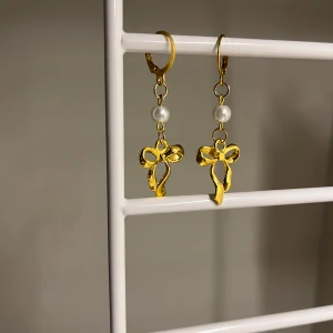 Guldiga örhängen med pärldetalj och rosett - Supersöta örhängen i guld med hängande rosett och en liten vit pärla ovanför. Perfekta för att ge din look en gullig och trendig touch. Lätta att bära och passar till många stilar. Själva ringen du har i örat är rostfritt stål. Handgjort 