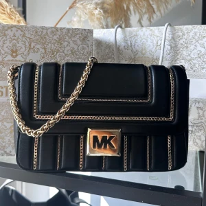 Svart Michael Kors axelväska i skinn - Snygg svart axelväska från Michael Kors i skinn med guldfärgade kedjedetaljer. Väskan har en justerbar axelrem i svart som medkommer. Väskan är köpt på Michael Kors i Göteborg 2022 & är en Limited edition utgåva. Det stora MK märkes på framsidan har lite slitningar (se bild).