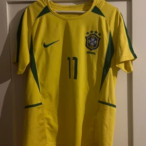 Brasilien 2002 Ronaldinho Nike fotbollströja M/L - Gul Brasilien fotbollströja från Nike med gröna detaljer, nummer 11 och Ronaldinho på ryggen. Klassisk rund hals och korta ärmar. Tillverkad i lätt och ventilerande polyester, perfekt för fotbollsälskare. Lite stor i storleken passar M/L 