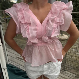 Rosa volangblus med v-ringning - Råka beställe två likadana, helt ny från förpackning. Supersöt ljusrosa blus med stora volanger framtill och v-ringning. Blusen har knappar framtill, ärmlös design med breda axelvolanger och smockad rygg. Tillverkad i ett tunt, transparent syntetmaterial som ger en drömmig look.