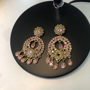 Guldiga örhängen med rosa stenar - Stora, statement örhängen i guldton med massor av glittrande rosa stenar. De har en rund, blomformad topp och hängande droppformade detaljer. Perfekta för att sticka ut och ge en lyxig känsla till din look.