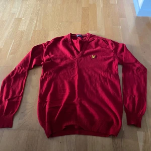 Röd v-ringad tröja från Lyle & Scott - Röd v-ringad tröja från Lyle & Scott Vintage med broderad gul logga på bröstet. Tröjan är långärmad och har ribbade muddar vid ärmslut och nederkant. Perfekt för dig som gillar klassisk stil med en färgstark touch.