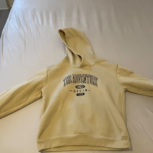 Beige hoodie med tryck THE ADVENTURE - Beige hoodie med huva och trycket 'THE ADVENTURE BEGIN 123' på bröstet. Mjuk insida och ribbade muddar vid ärmslut och nederkant. Perfekt för chill dagar eller när du vill ha en avslappnad look.