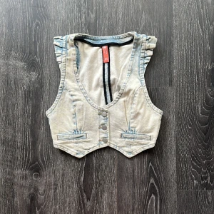 Ljus jeansväst med volangdetaljer - Snygg ljusblå jeansväst med volangdetaljer på axlarna och markerad midja. Västen har djup v-ringning, tre knappar framtill och dekorativa fickor. Perfekt att styla över en t-shirt eller klänning för en cool look.