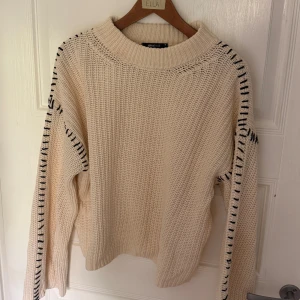 Beige stickad tröja från Gina Tricot  - Säljer en beige stickad tröja från Gina tricot med svarta dekorativa sömmar längs ärmarna. Tröjan har en chunky känsla, rund halsringning och långa ärmar. Perfekt för lager-på-lager och chill höststil.