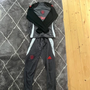Manchester United träningsset Adidas - Snyggt Manchester United träningsset från Adidas med långärmad tröja och byxor. Tröjan är grå med svarta ärmar, röda detaljer och klubbmärke samt Tezos-logga på bröstet. Tillverkad i lätt och ventilerande material (Aeroready) perfekt för träning.