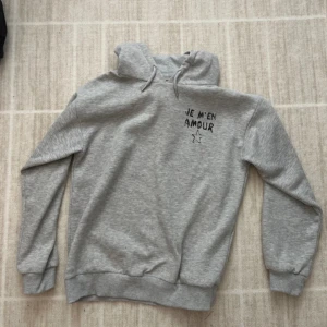Grå hoodie med tryck Je m'en amour - Säljer en grå hoodie med svart tryck 'Je m'en amour' och en stjärna både fram och bak. Tröjan har huva med snörning och ribbade muddar vid ärmslut och nederkant.