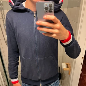 Moncler hoodie  - Snygg marinblå hoodie från Moncler med hel dragkedja, huva och fickor framtill. Ärmsluten har röd, vit och blå randig mudd. Moncler-logga på vänster ärm och coola detaljer på huvan. Perfekt för dig som gillar streetwear och vill ha något stilrent men ändå sportigt.