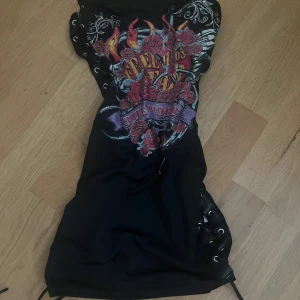 Svart kort klänning med snörning - Svart kort klänning med coolt tryck framtill i rött, lila och gult med rosor och flames. Snygga snörningsdetaljer längs båda sidorna ger en edgy vibe. Perfekt för dig som gillar statement-plagg och rockig stil.