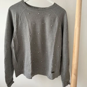 Grå sweatshirt med pärlor Abercrombie - Grå sweatshirt från Abercrombie & Fitch med snygga pärldetaljer framtill. Tröjan har rund hals, långa ärmar och en relaxed passform. Perfekt för dig som gillar en enkel men ändå unik stil. Mjuk bomullskänsla och diskret logga vid nederkanten.