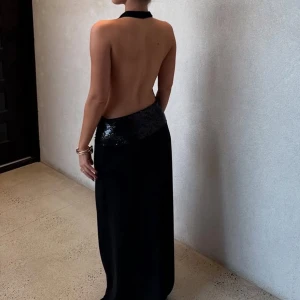 Svart långklänning med paljettdetalj - Snygg svart långklänning med halterneck och öppen rygg. Klänningen har en glittrig paljettpanel i midjan som ger extra edge. Modellen är ärmlös och har en elegant, slank siluett. Perfekt för dig som vill sticka ut med en stilren och glam look.