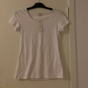 Vit basic t-shirt från Lindex - En klassisk vit t-shirt från Lindex Essentials med rund halsringning och korta ärmar. Gjord i mjuk bomull som känns skön mot huden. Perfekt att styla med jeans eller kjol för en clean och enkel look.