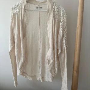 Beige kofta med spets från Abercrombie - Luftig beige kofta från Abercrombie & Fitch med långa ärmar och snygga spetsdetaljer på axlarna. Stickad i ett tunt material. Perfekt att slänga över en topp när det är lite kyligt ute.