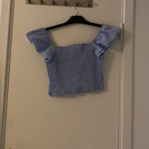 Ljusblå croppad blus med volangärmar - Supersöt ljusblå croppad blus med fyrkantig urringning och volangiga korta ärmar. Toppen har smockad framsida och slät baksida, vilket ger en snygg passform. Perfekt för dig som vill ha en trendig och luftig topp till sommaren.